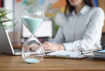 Glass hourglass standing against background of woman working at laptop closeup. Working hours concept